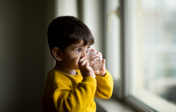 Child looking out of a window, drinking water from a glass.