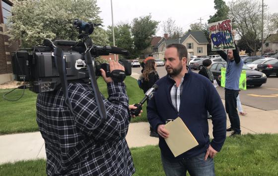 MI - Sean McBrearty being interviewed by TV crew with protestors in background.